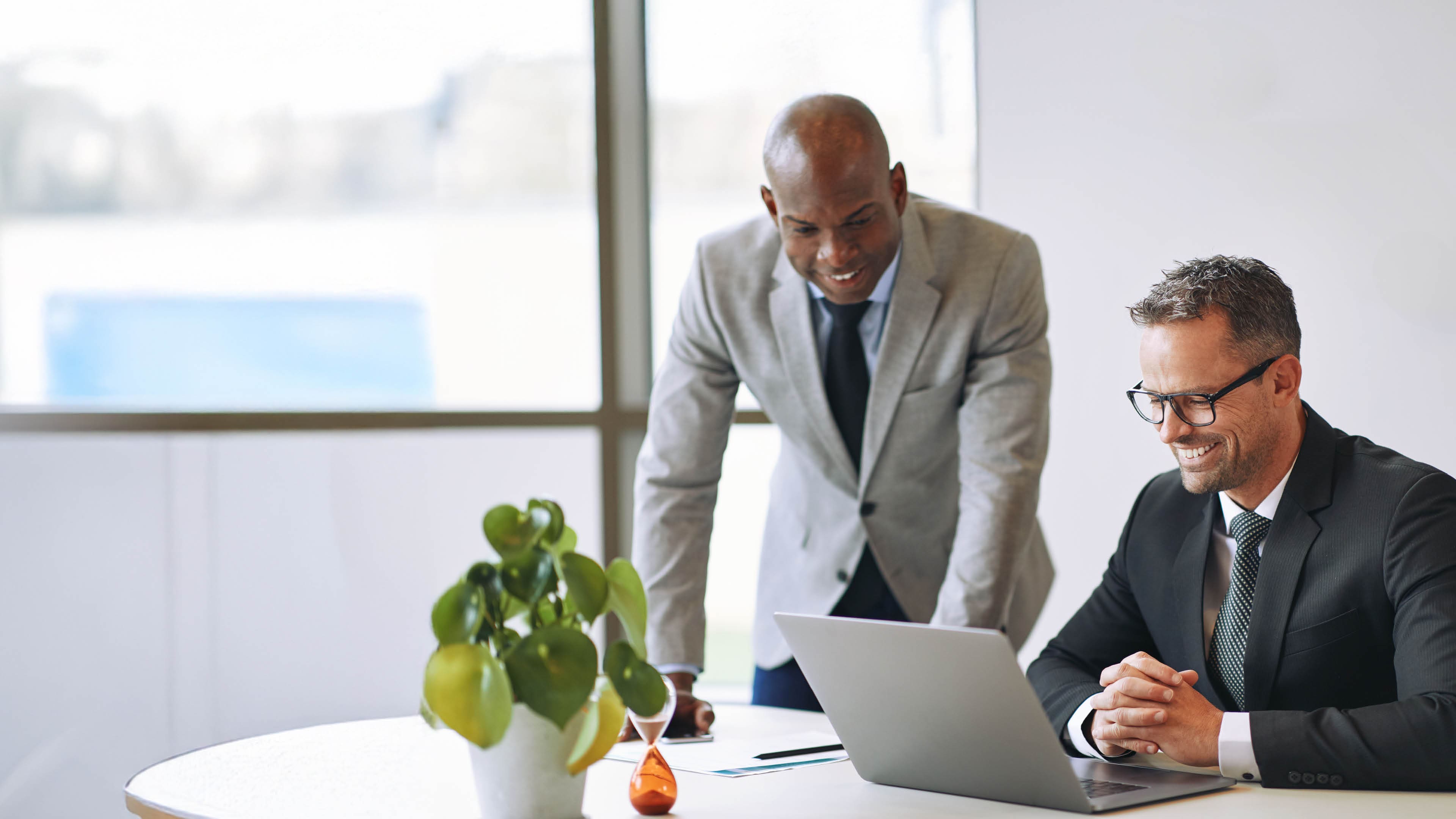 Two diverse businessmen smiling and talking together while working on a laptop at a table in an office